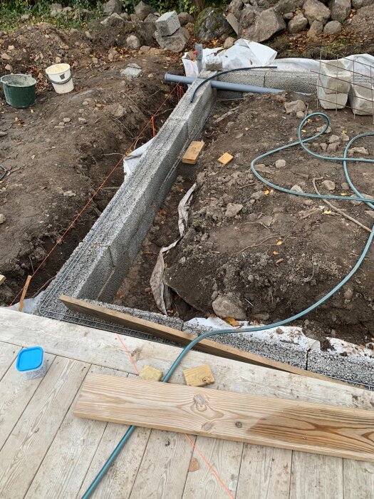 Stenmurkonstruktion med betongblock och stenar på en byggarbetsplats, uppmärkta linjer och verktyg synliga på marken.