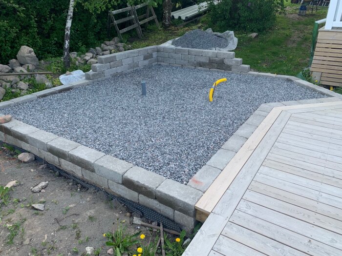 Stenterass med stödmur och stenkross på sluttande tomt, omgiven av träd och trätrall; gul slang och rör synliga.