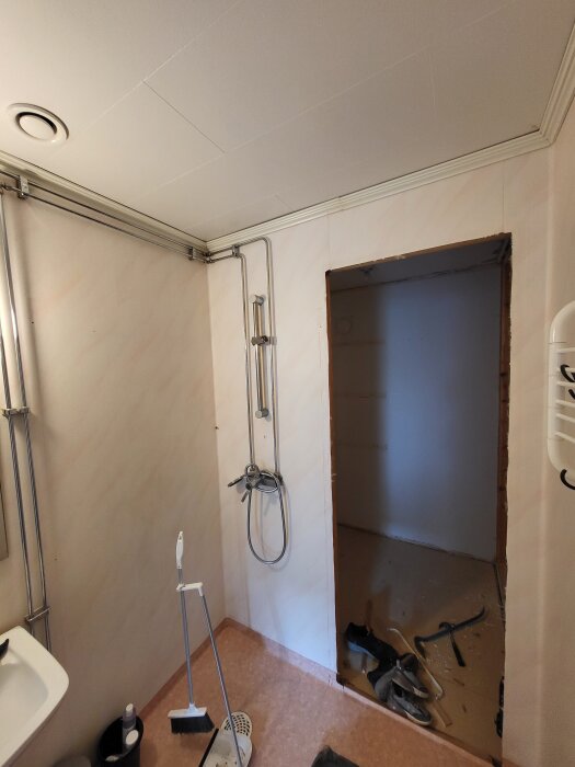 Badrum under renovering med dusch, verktyg på golvet och oavslutad dörröppning till ett annat rum. Frånluftsventil synlig i taket.