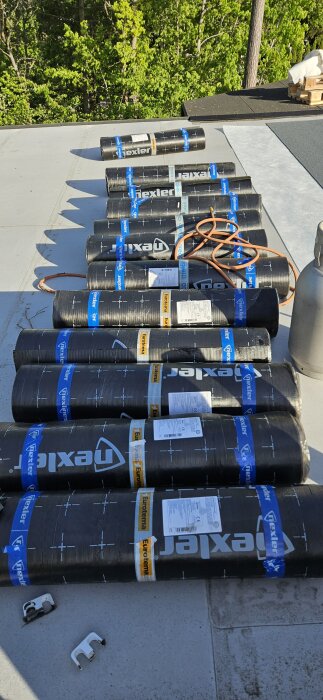 Rullar av Nexler Izolmat Plan monomax takmaterial ligger utbredda på ett tak i en grönskande omgivning.