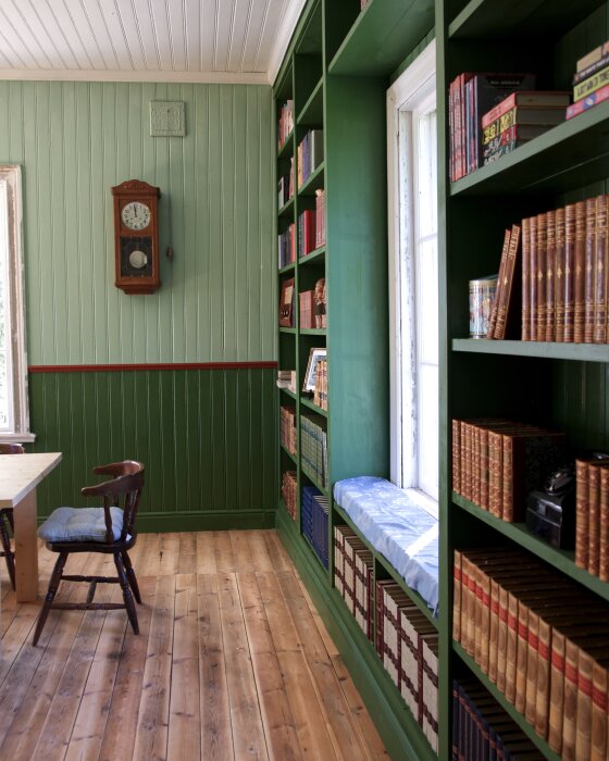 Old schoolhouse interior with wooden floor, green bookshelves, a wall clock, and a small wooden chair by a table, creating a cozy reading nook.