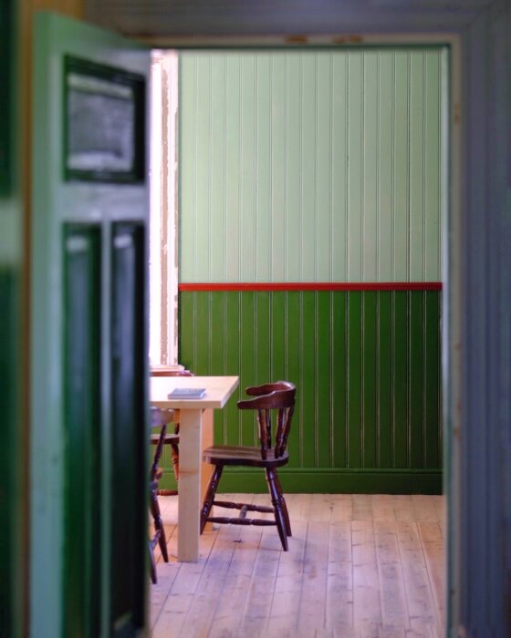Öppet dörrhörn visar rum med grön träpanel, ett träbord och en stol, vilket framhäver husets renoveringsstil.