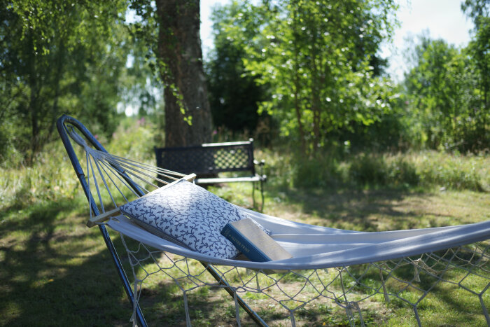 Hängmatta med kudde och bok i trädgårdsmiljö, omgiven av träd och en bänk i bakgrunden.
