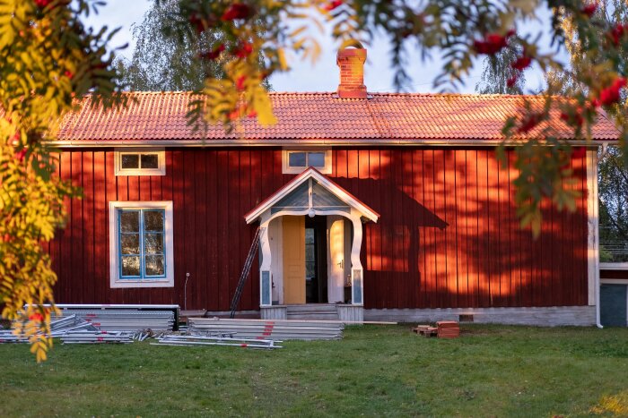 Rött gammaldags skolhus med tegeltak, omgiven av träd och byggmaterial vid entrén.