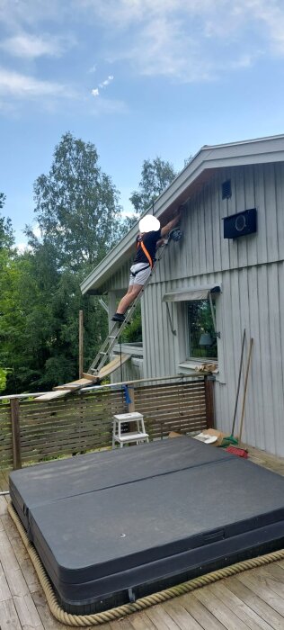 Person klättrar på stege, utför arbete på takfot av hus över en altan, med grönt landskap i bakgrunden.