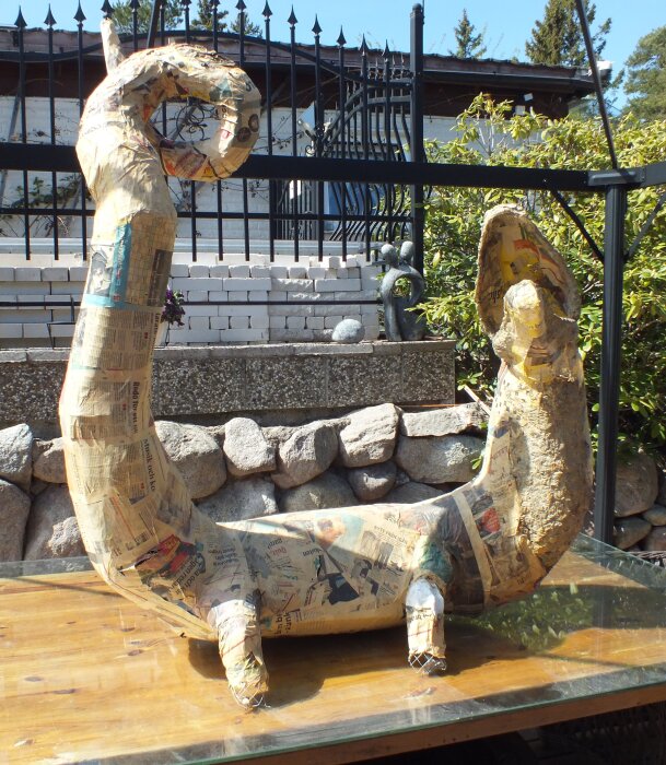 Skulptur av en sittande drake gjord av hönsnät och täckt med papper, placerad utomhus på ett träbord, med stengärdsgård i bakgrunden.