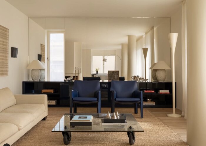 Elegant vardagsrum med spegelvägg, blå stolar, beige soffa och glasbord, reflekterande dagsljus och moderna inredningsdetaljer.