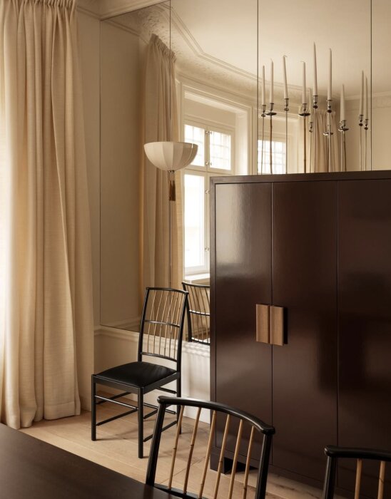 Elegant rum med stora speglar som täcker en vägg, reflekterande ljus, ett mörkt träskåp och stolar med ljusa gardiner vid ett fönster.
