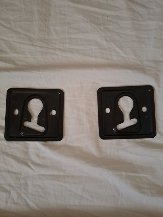 Två svarta väggfästen för TV-montage, märkta med L och R, ligger på en vit yta.