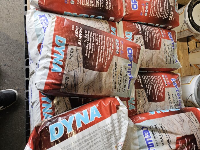 Flera påsar med Dyna polymeric sand i vitt och rött, märkta med "Granite Grey", ligger staplade på varandra i en stenbutik.