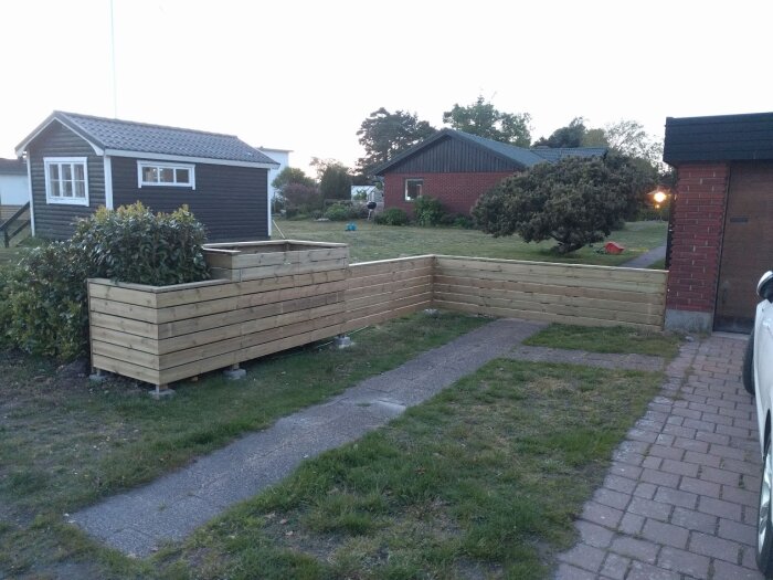 Träinramade avfallslådor för fyrfackssystem bredvid en trädgårdsgång, med buskar och hus i bakgrunden.