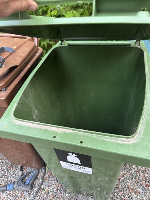 Grön soptunna öppen med etikett märkt "restavfall", hand syns vid locket, traditionell brun soptunna i bakgrunden, grus under.