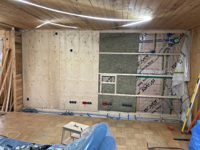 Rum under renovering med träpanel på vägg och tak, inspektionsöppningar, isolering och delvis exponerad rördragning. Parkettgolv med verktyg och en stege.