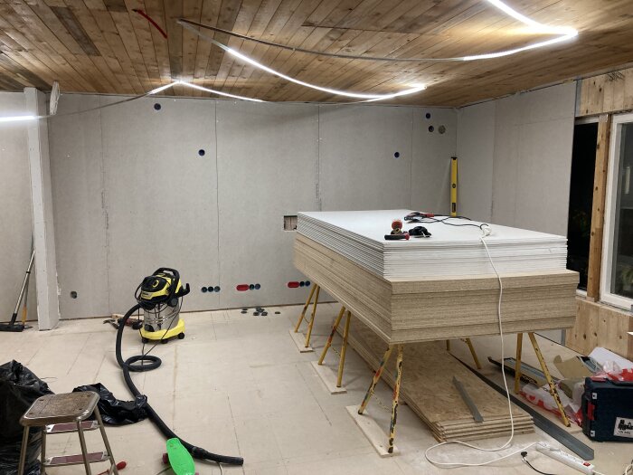 Renoveringsrum med staplade gipsskivor, en dammsugare och verktyg på ett bord.