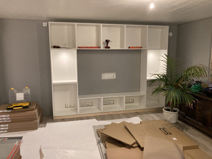 Vit bokhylla under montering med verktyg och kartonger runtomkring; växt vid sidan.