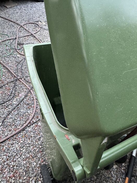 Grönt avfallskärl som lutas för tömning på en grusig yta, omgiven av trädgårdsslangar i bakgrunden.