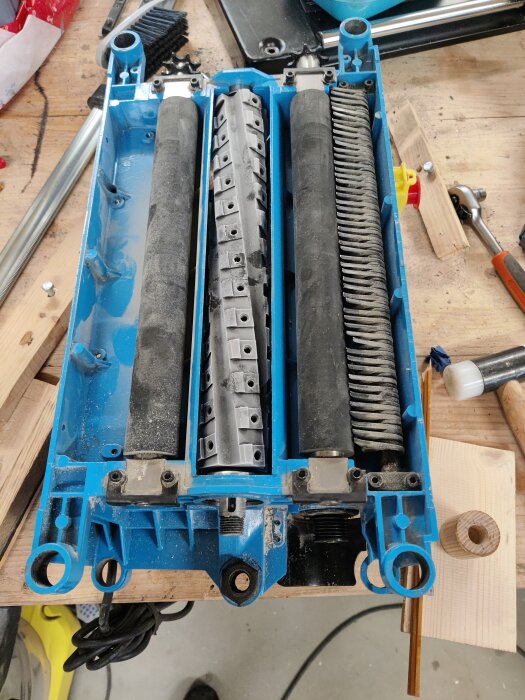 Bild på en isärtagna planhyvel med monterad helix-cutter och två valsar, belägen på en arbetsbänk tillsammans med verktyg och träkomponenter.