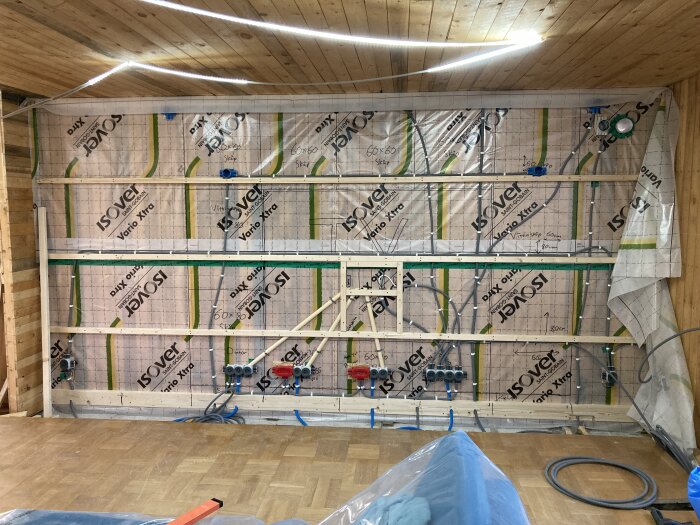 Vägg under konstruktion med träreglar och plastisolering från Isover. Synliga elledningar och rör förbereder för kommande installationer.