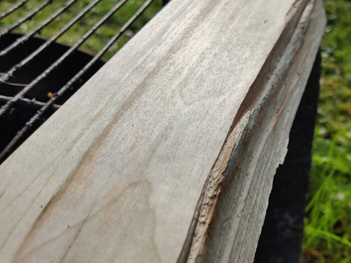 Närbild av en träbit med slät yta, troligen från en planhyvel, mot en bakgrund av metallgaller och gräs.