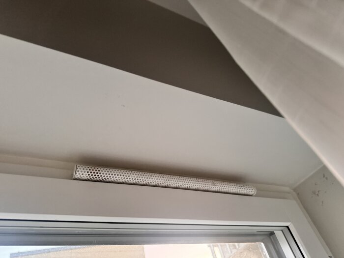 Ventilationsöppning ovanför fönster med synlig gardin, där kall luft drar in i ett rum.