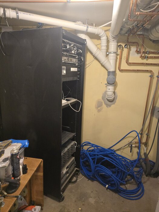 Serverrack i källare med patchkablar och rörledningar på väggarna. Verktyg och tillbehör syns på en träbänk framför.