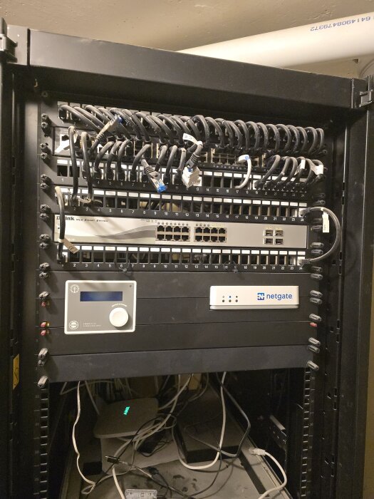 Serverrack med nätverksswitch, kablar och Netgate-enhet i ett hobbyrum, omgivna av interna nätverksanslutningar och IT-utrustning.
