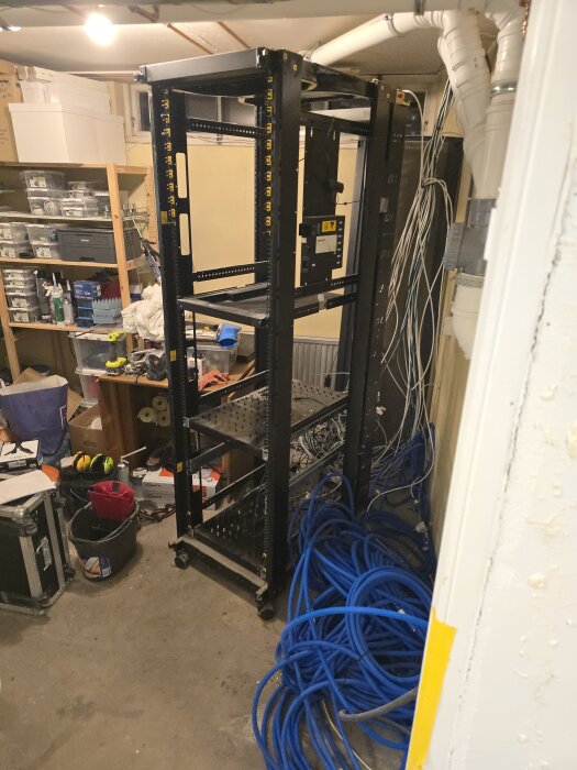 Ett tomt, svart serverrack står i ett rum med många kablar och utrustning runtomkring. Det finns kabelhärvor på golvet.