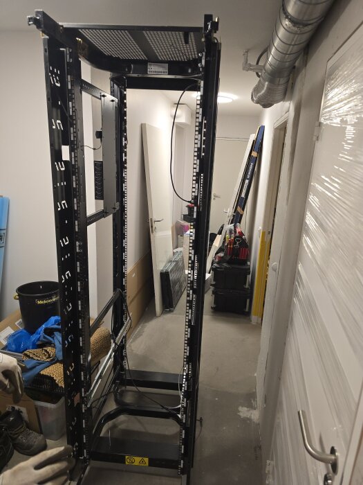 Serverrack utan sidoplåtar i en trång korridor, omgiven av verktyg och flyttkartonger. Racket är redo att monteras i ett maskinrum.