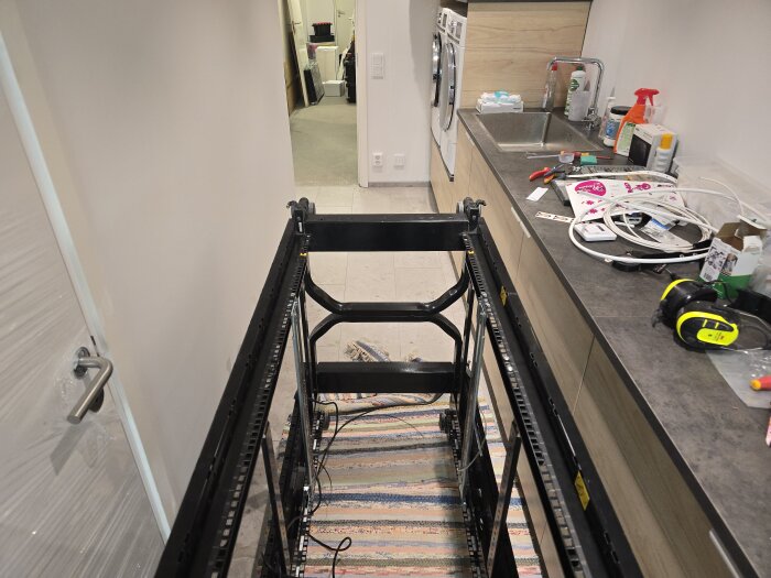 Ett serverrack som ligger på trasmattor i en tvättstuga, omgiven av verktyg och kablar. Väggen har en dörr och en bänkskiva med diskho.