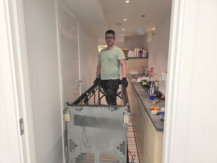 Man i t-shirt och handskar flyttar ett stort serverrack genom en smal korridor, omgiven av verktyg och förpackningar.