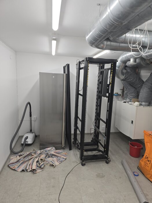 Tomt serverrack i rum med ventilationsrör, dammsugare och trasmatta på golvet.