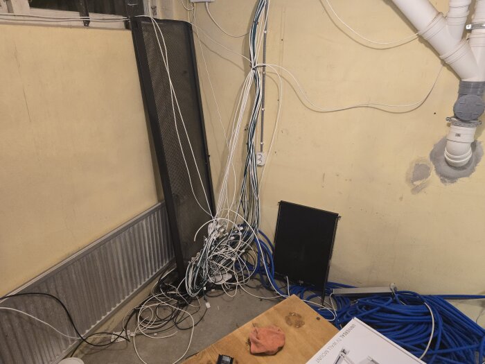 Ett hörn av ett rum med en hög av kablar på golvet, en svart serverrack-dörr lutad mot väggen och synliga ventilationsrör.