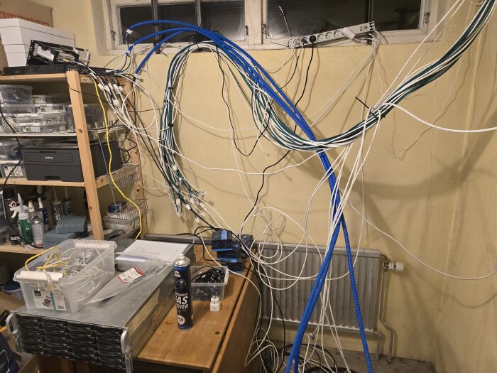 Kabelkaos i hobbyrum med nätverkskablage, verktyg, och hyllsystem. Serverutrustning och diverse snickeritillbehör syns i rörig miljö.