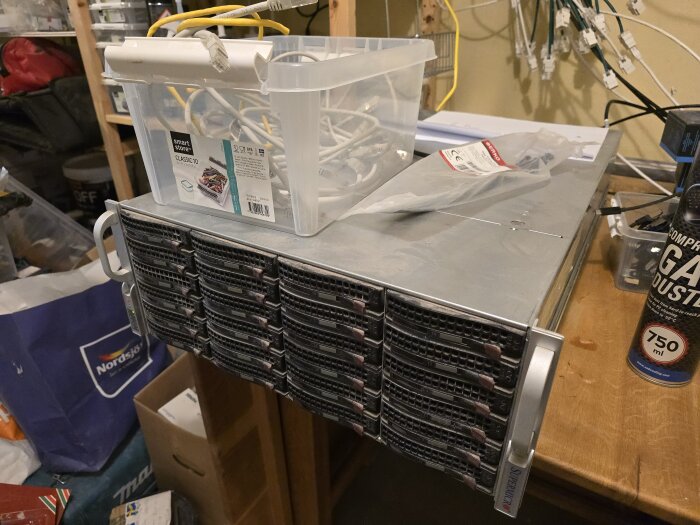Dammig 4U-server med många diskar ovanpå ett träbord, plastlåda med kablar och etikett "SmartStore Classic 10", komprimerad gasburk synlig.