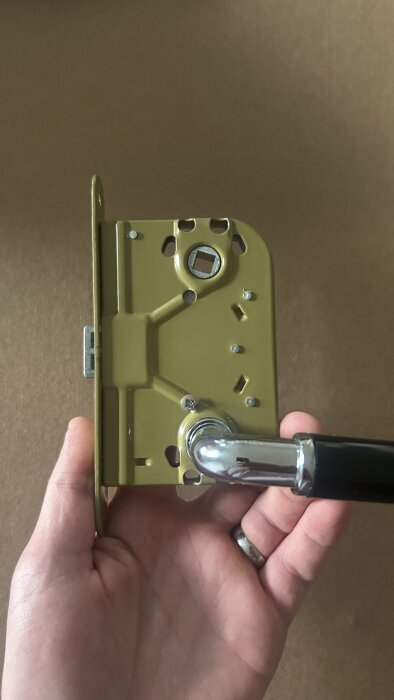 En hand håller en metalldel av ett lås, visas från sidan med och utan skruv monterad.