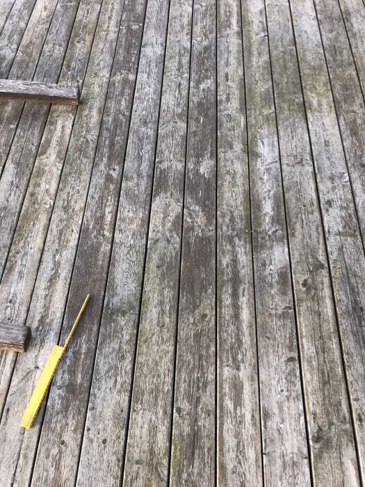 Sliten trall med ytskydd som lossnat, visar tecken på alger och svartmögel. En gul pensel ligger på träplankorna.