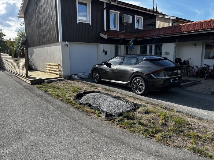 Altan och staket vid ett hus med makadam och svart bil på uppfarten; gräsmatta med grus i förgrunden, ska enligt plan asfalteras eller stensättas.