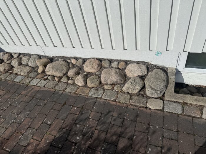 Stora stenar placerade som dränering bredvid en husvägg, intill en stenlagd gång.