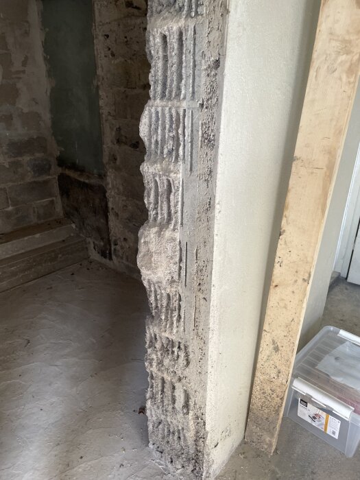 Rough stone pillar in a basement, adjacent to a plastered wall and next to a plastic storage box.