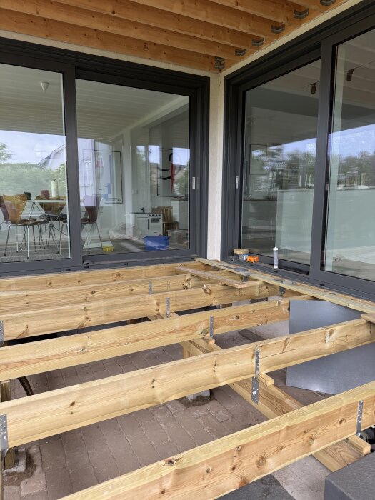 Trädäck under konstruktion framför glaspartier i en hörnvinkel, träreglar på plats, inredning och trädgård synlig genom glaset.