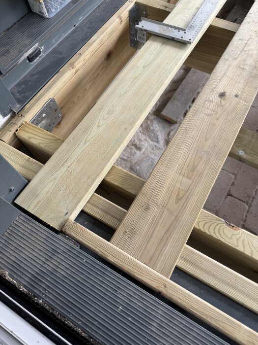 Trädäck under konstruktion med träbalkar och metallbeslag nära en skjutdörr av glas. En snickarvinkel ligger på en bräda.