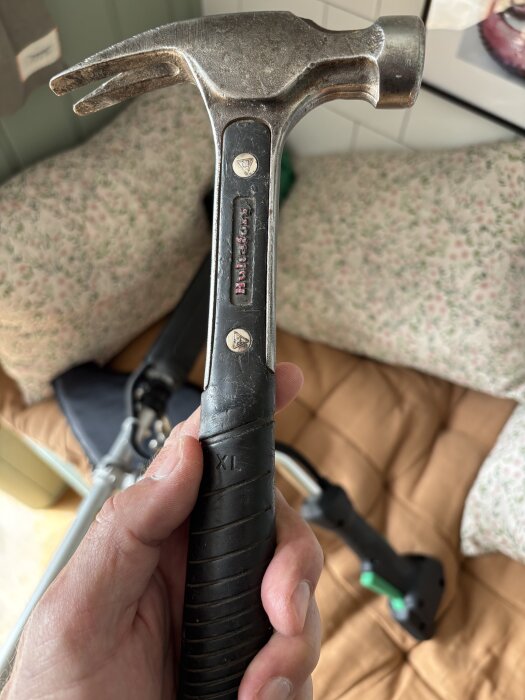 En nära bild på en hammare med svart gummihandtag och metallhuvud, som hålls i en hand ovanför en mysig filt.