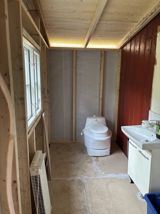 Tillbyggnad under konstruktion med träram, fönster och isolering; ett provisoriskt badrum med toalett och handfat.