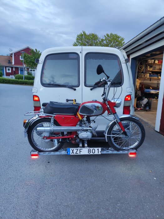 Röd moped monterad på dragkrok bakom vit skåpbil, redo för mopedrally och camping.