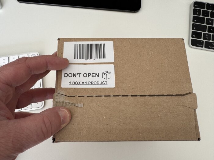 Kartong med etikett "DON'T OPEN" och streckkod, hålls av en hand. I bakgrunden syns två tangentbord.