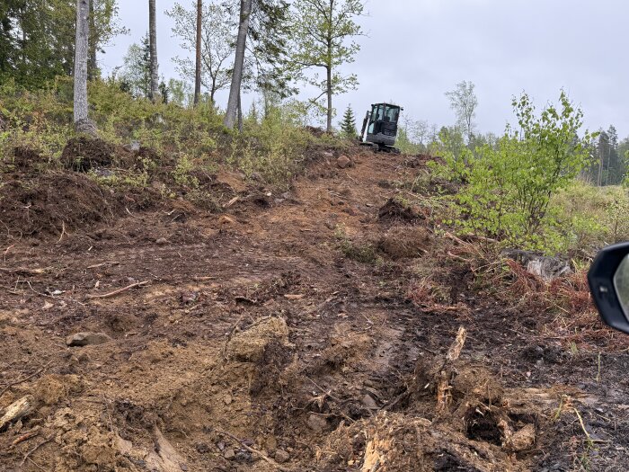 Grävskopa på en lerig och kuperad skogsterräng omgiven av träd och buskar, med grå himmel i bakgrunden.