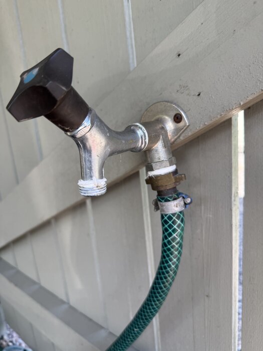 Utvändig vattenkran med mörkt handtag och en ansluten grön slang, monterad på en trävägg. Gängad del synlig på kranen.