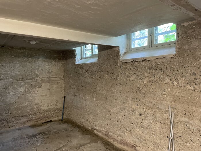 Grov källarvägg av betong med småsten, rester av gammal puts. Två fönster upptill, lite byggverktyg på golvet. Naturligt ljus från fönstren.