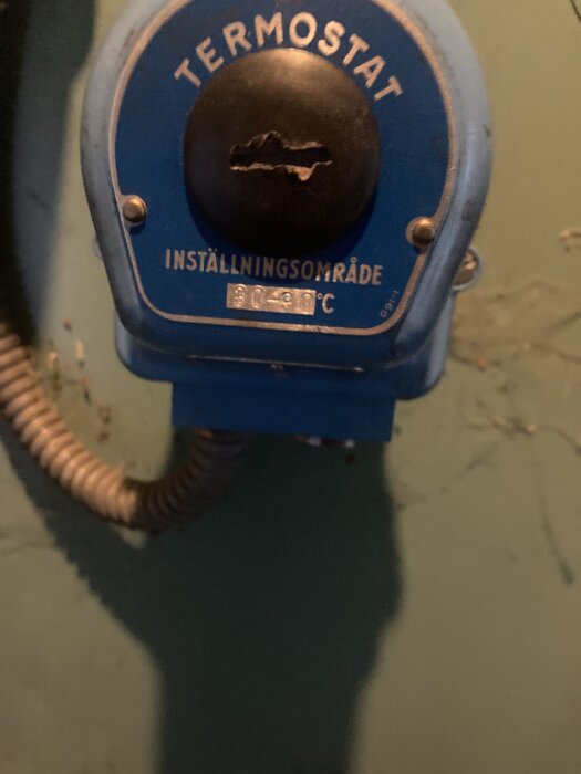Blå termostat med justeringsområde 30-80°C, monterad på en vägg intill en metallslang.