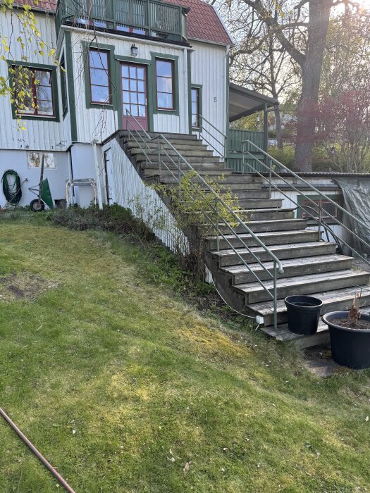Gammal stentrappa med metallräcke utanför ett hus med vita väggar och rött tak, omgiven av gräsmatta och träd.